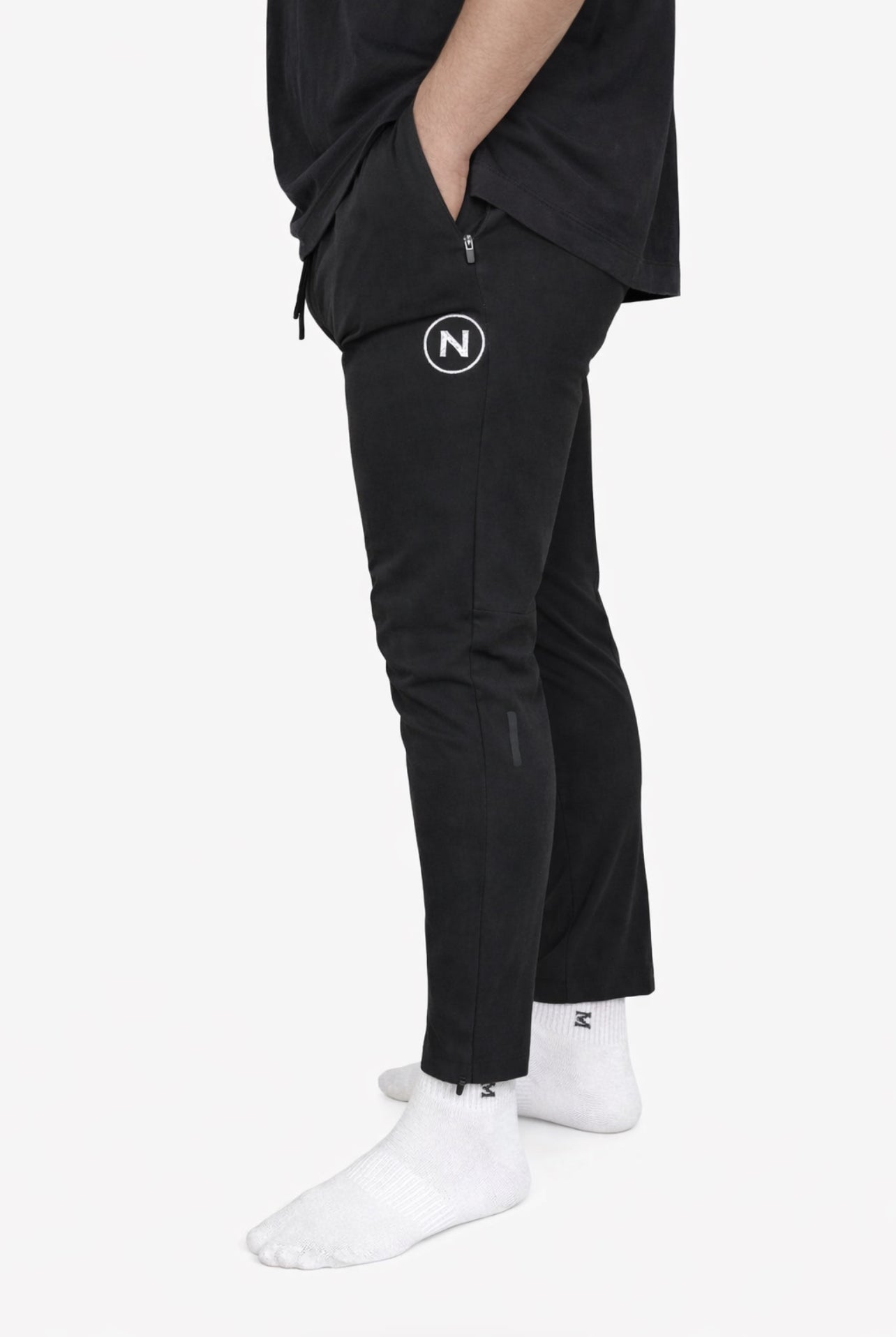 PRONOAM black sports trousers against a white background. The pants feature a circular "N" logo on the upper thigh, a zippered side pocket, and a tapered fit that ends at the ankle, paired with white socks.